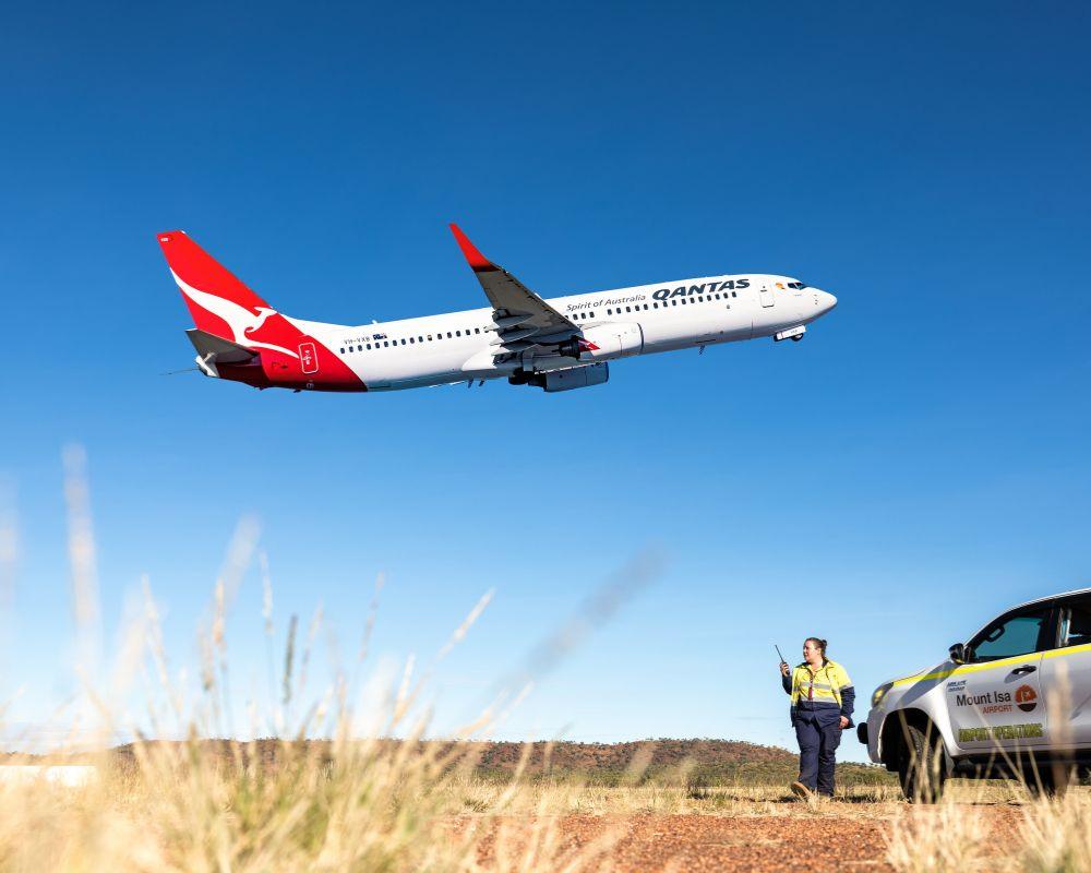 Qantas airplane taking off at Mount Isa Airport with an airport worker and vehicle in the foreground.