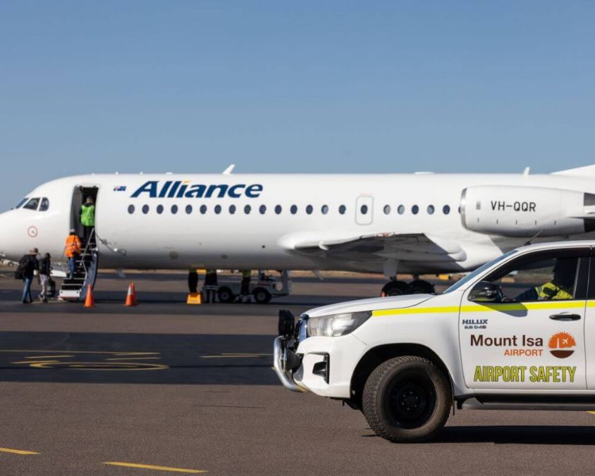 Alliance Airlines aircraft VH-QQR at Mount Isa Airport with passengers boarding and an airport safety vehicle in the foreground.