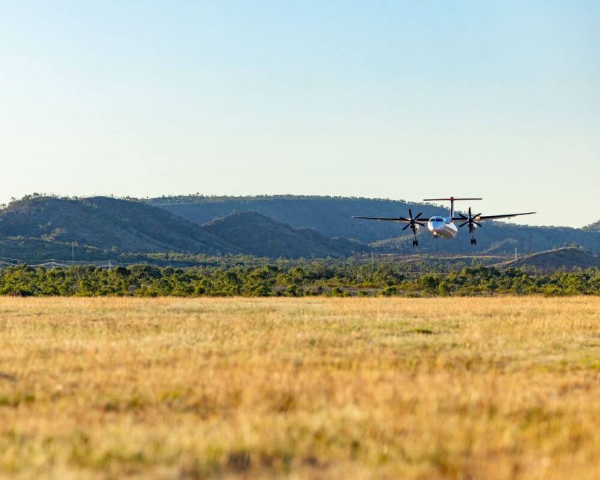 Propeller aircraft flying low over a grassy field near Mount Isa, with hills and clear sky in the background.