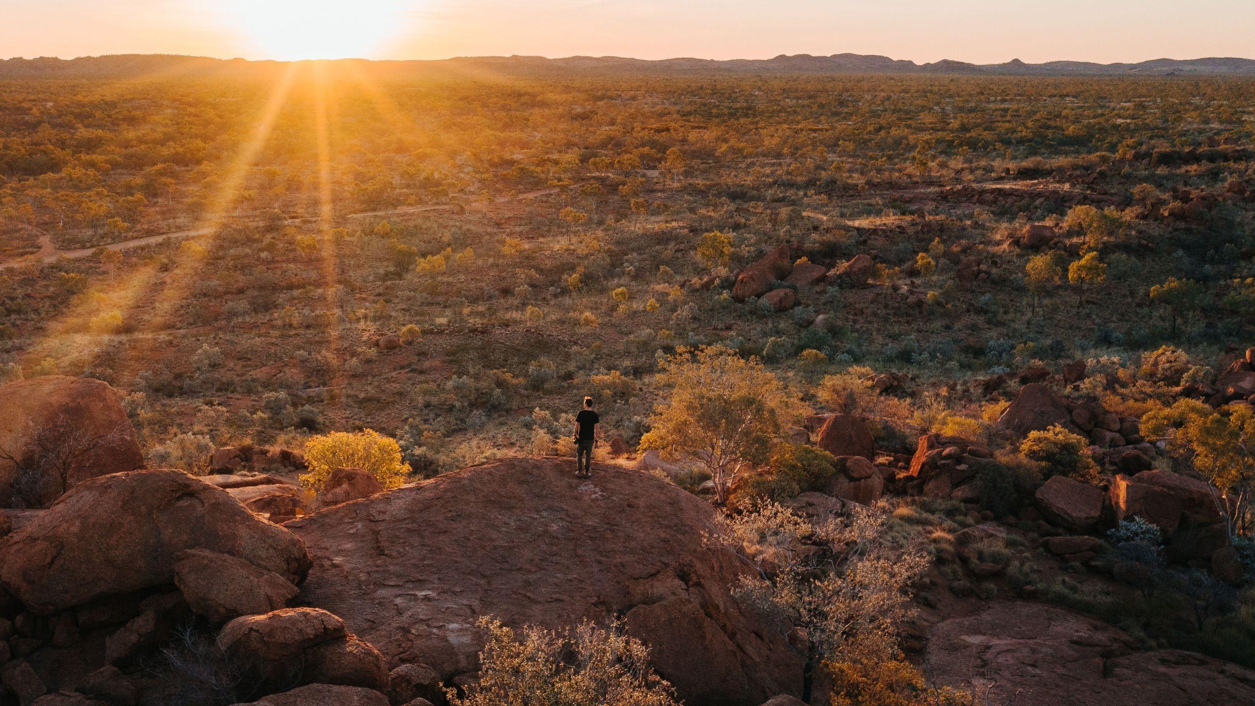 Person standing on a rocky outcrop overlooking the rugged outback landscape of Mount Isa at sunset, with golden light illuminating distant hills and sparse vegetation.