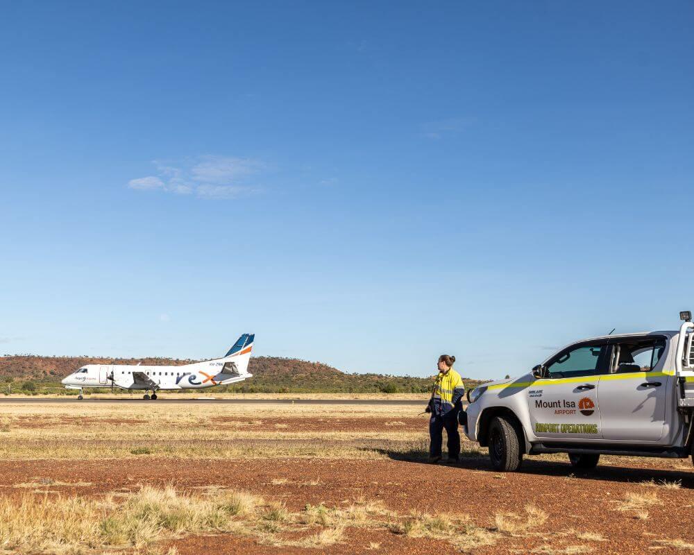 Regional Express (Rex) aircraft on the runway at Mount Isa Airport with a utility vehicle and airport staff member in high visibility clothing nearby.