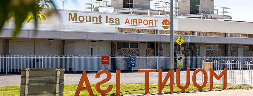 Exterior view of Mount Isa Airport featuring large orange “MOUNT ISA” signage, airport entrance, and directional signs for parking and pedestrian crossing.