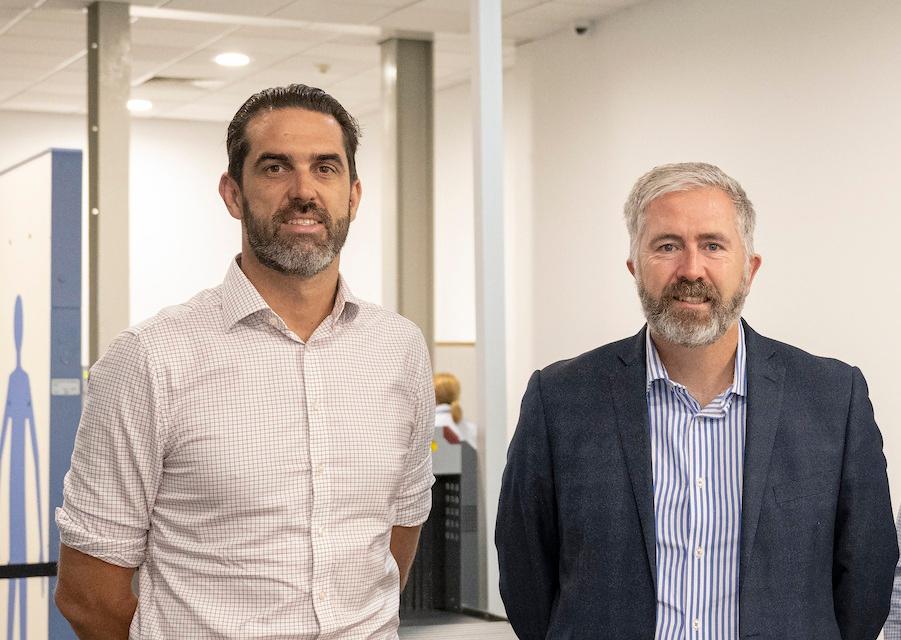 Two QAL employees standing indoors at Mount Isa Airport, one in a checkered shirt and the other in a blazer, with office-style pillars and ceiling lights in the background.