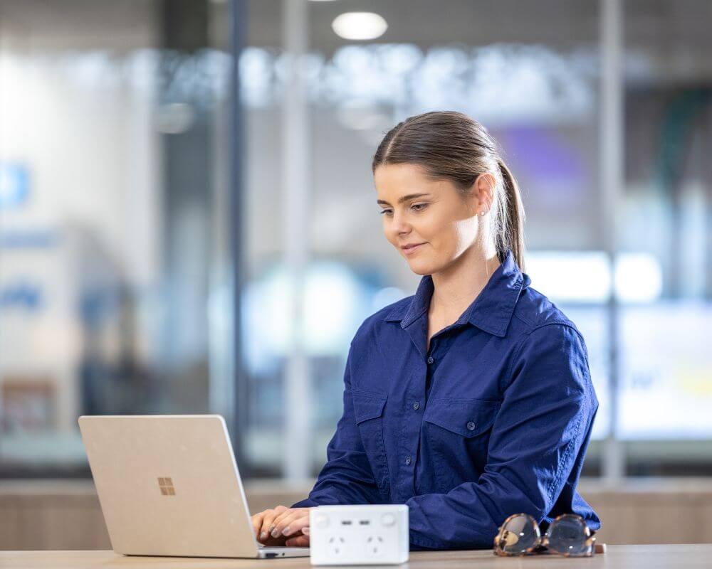 Person in a blue shirt working on a laptop at Mount Isa Airport, with sunglasses and a power hub on the desk in a modern office setting.