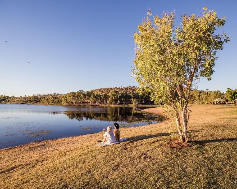 Two people sitting on a blanket under a tree by a lake near Mount Isa, with reflections of the sky and surrounding greenery in the water.