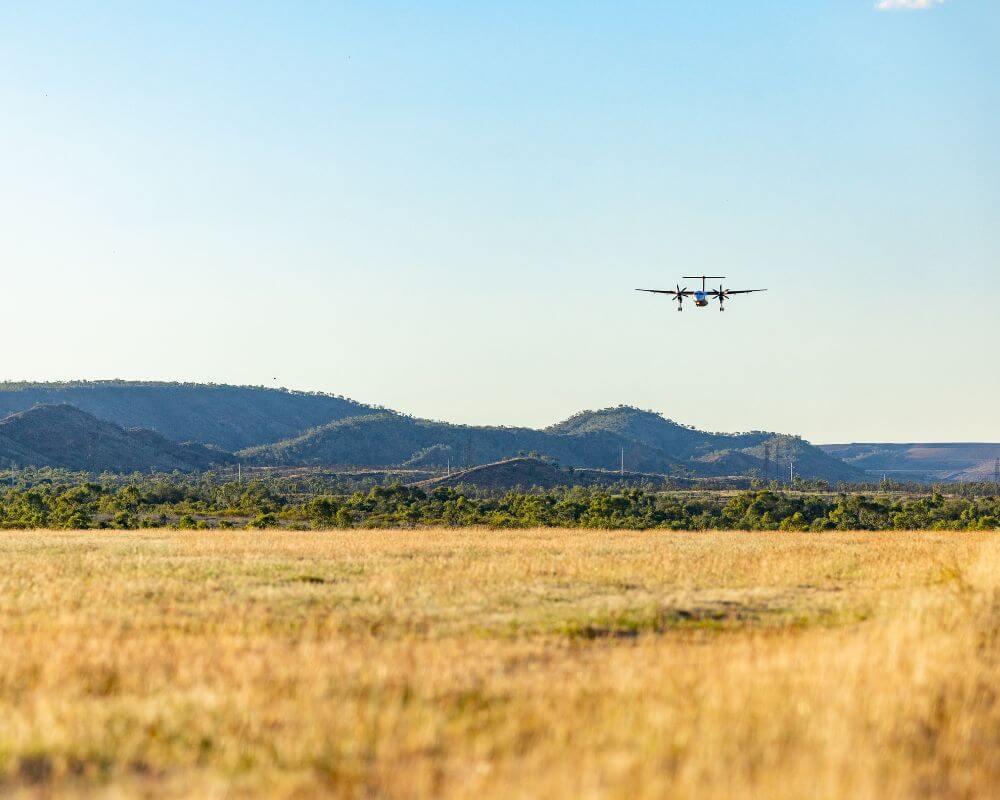 Small aircraft landing or taking off over grassy terrain with Mount Isa’s hills and clear blue sky in the background.