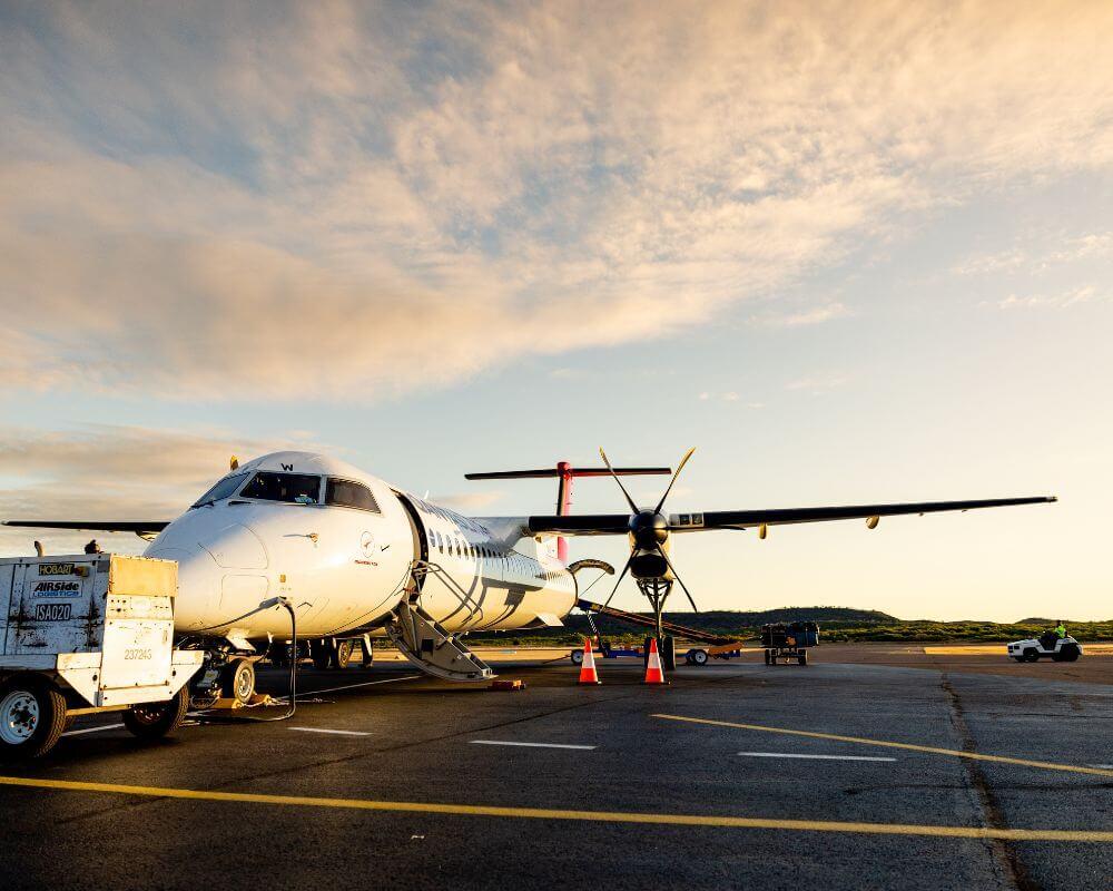 Propeller aircraft parked on the tarmac at Mount Isa Airport during sunrise or sunset, with orange cones and a ground service vehicle nearby.