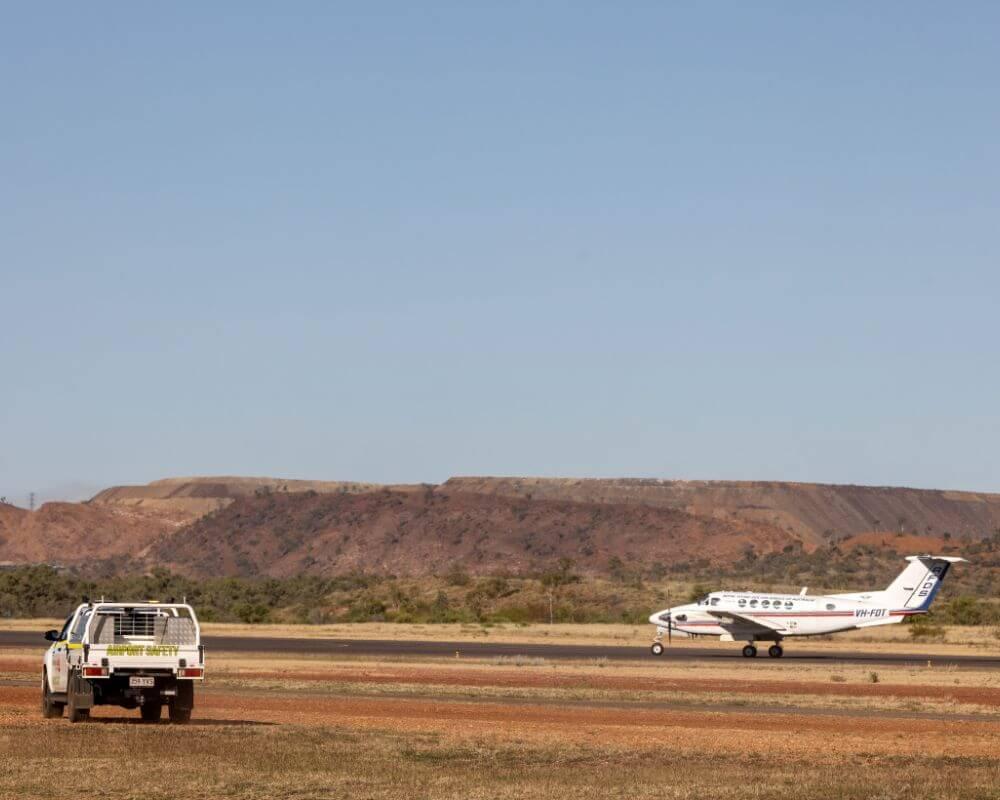 Small aircraft VH-FXT on the runway at Mount Isa Airport with a white utility vehicle nearby and hills in the background under a clear blue sky.