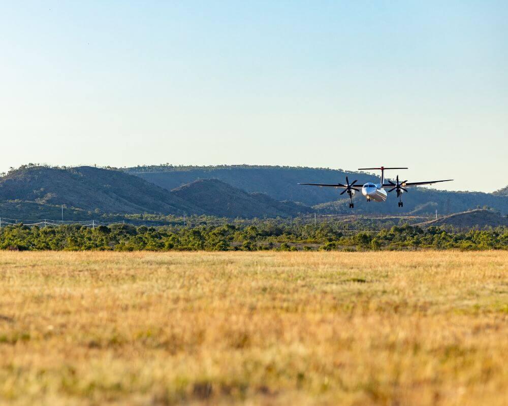 Propeller aircraft flying low over a grassy field near Mount Isa, with hills and clear sky in the background.