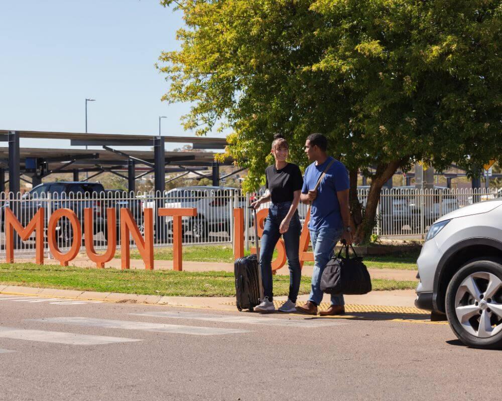 Two people walking with luggage past a large 'Mount Isa' sign near the airport parking area, with vehicles and a shaded tree in the background.