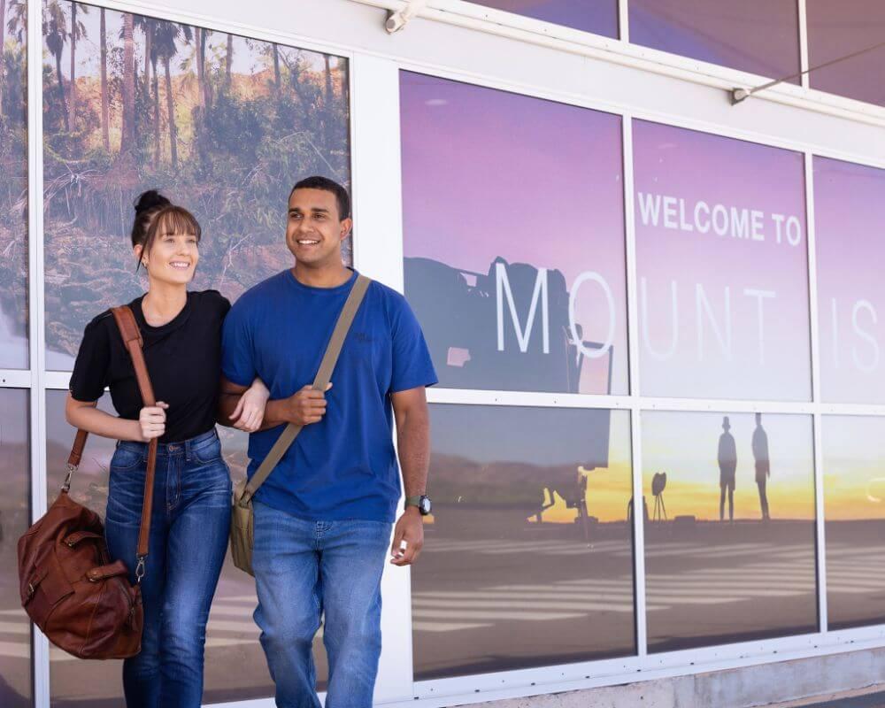 Two people walking arm in arm with bags in front of Mount Isa Airport, passing a large 'Welcome to Mount Isa' sign displayed on the terminal windows