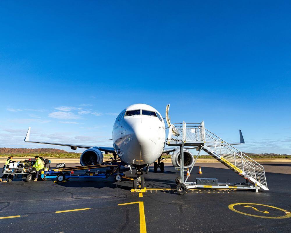 Commercial aircraft on the tarmac at Mount Isa Airport, with ground crew handling baggage and a mobile staircase positioned for boarding under a clear blue sky.