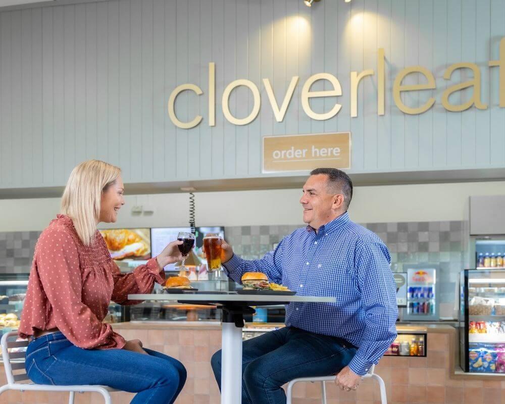 Two people toasting drinks at a high-top table inside Cloverleaf café, with sandwiches, fries, and a casual dining atmosphere featuring food displays and signage.