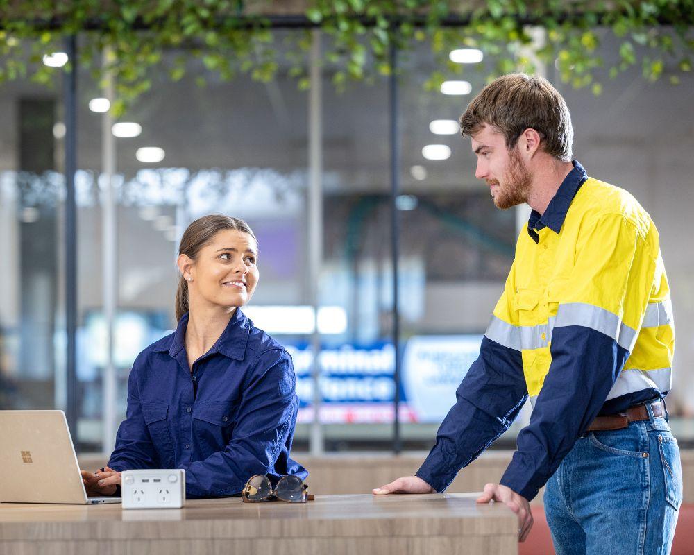 Two individuals interacting at a desk in the terminal of Mount Isa Airport, with a laptop and greenery visible in a modern office-style environment.