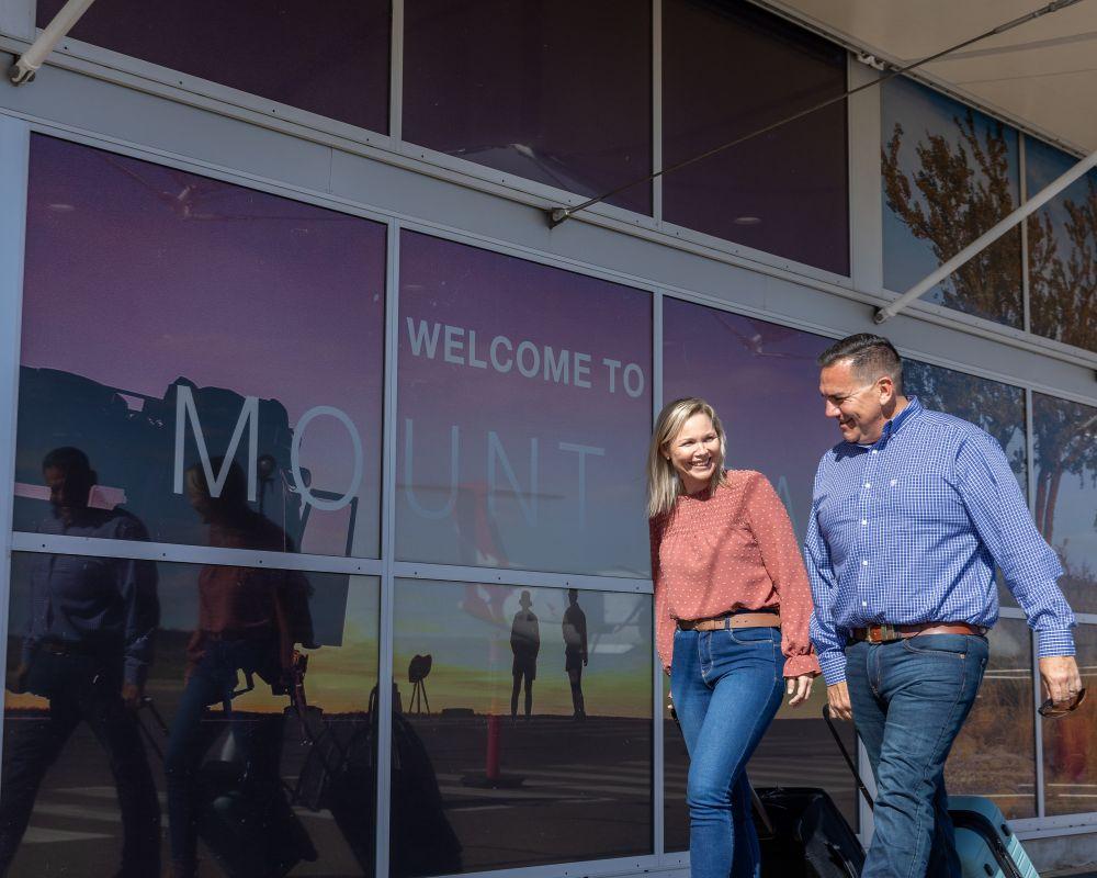 Two people walking outside the Mount Isa Airport terminal, with a large 'Welcome to Mount Isa' sign visible on the glass façade.