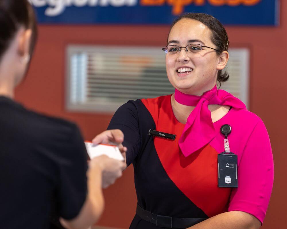 Staff member handing an item to a visitor in a customer service area inside Mount Isa Airport, with partial signage reading 'Regional Express' in the background.
