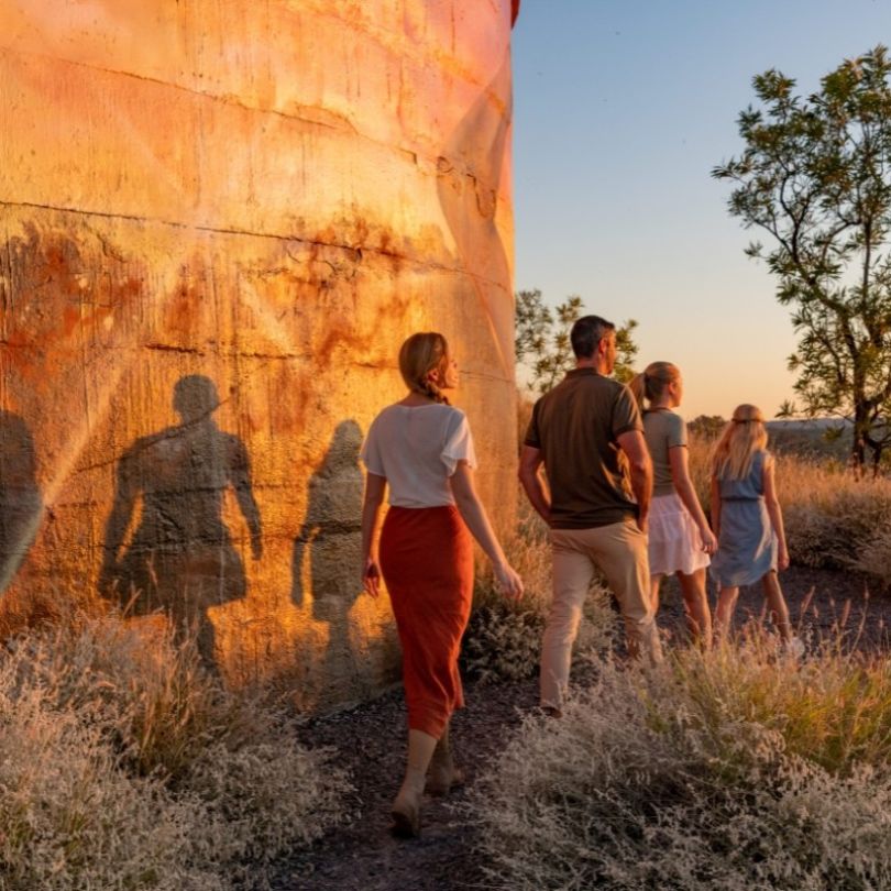 Visitors walking beside the Cloncurry Water Tank Mural at sunset, with long shadows cast on the cylindrical structure and outback scenery in the background.