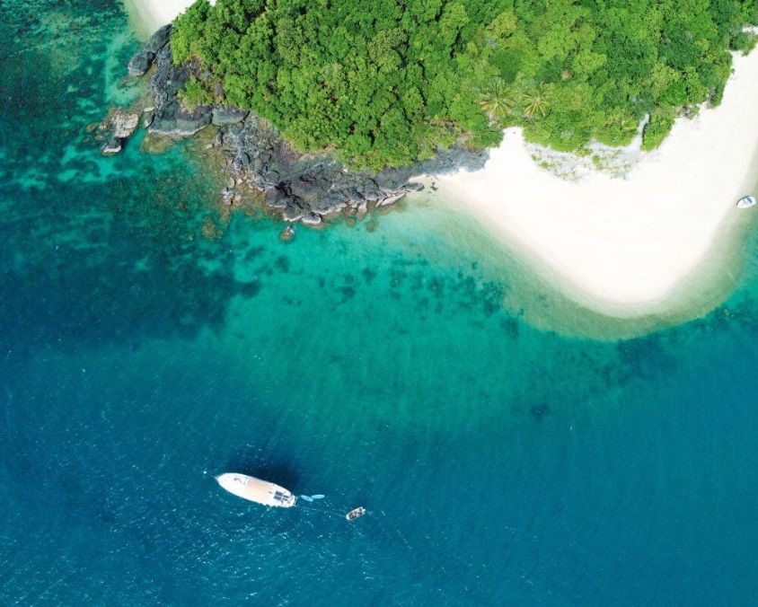Aerial view of a tropical island with lush green foliage, white sandy beach, and clear turquoise water. A boat floats just offshore, surrounded by vibrant blue ocean and coral reefs.