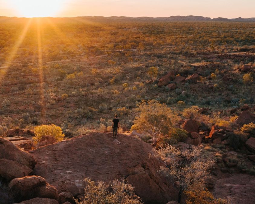 Person standing on a rocky outcrop overlooking the rugged outback landscape of Mount Isa at sunset, with golden light illuminating distant hills and sparse vegetation.
