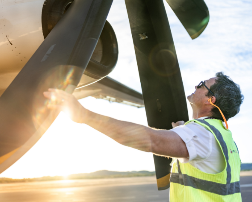 Ground crew member in high-vis vest inspecting an aircraft propeller at sunrise on the tarmac.