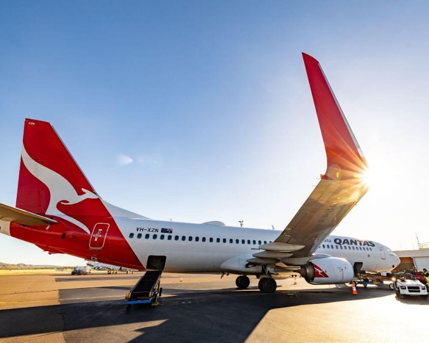 Qantas aircraft VH-XZN on the tarmac at Mount Isa Airport with ground service vehicles and personnel nearby under bright sunlight.