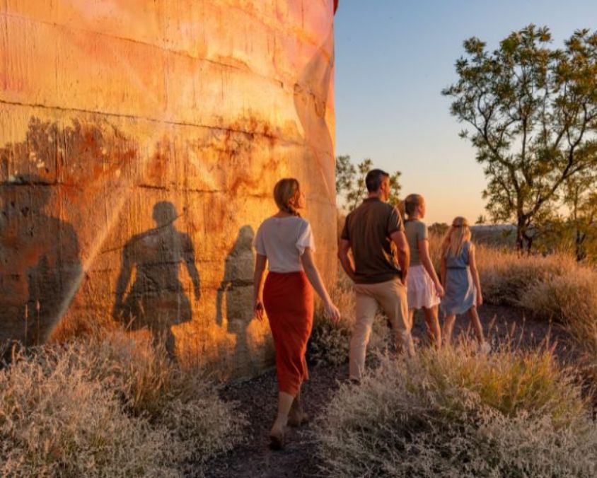 Visitors walking beside the Cloncurry Water Tank Mural at sunset, with long shadows cast on the cylindrical structure and outback scenery in the background.