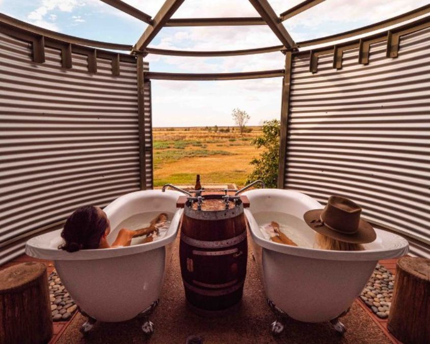 Two visitors soaking in outdoor clawfoot bathtubs at Julia Creek, with a scenic rural landscape in the background and a rustic setup featuring a shared wooden barrel faucet.