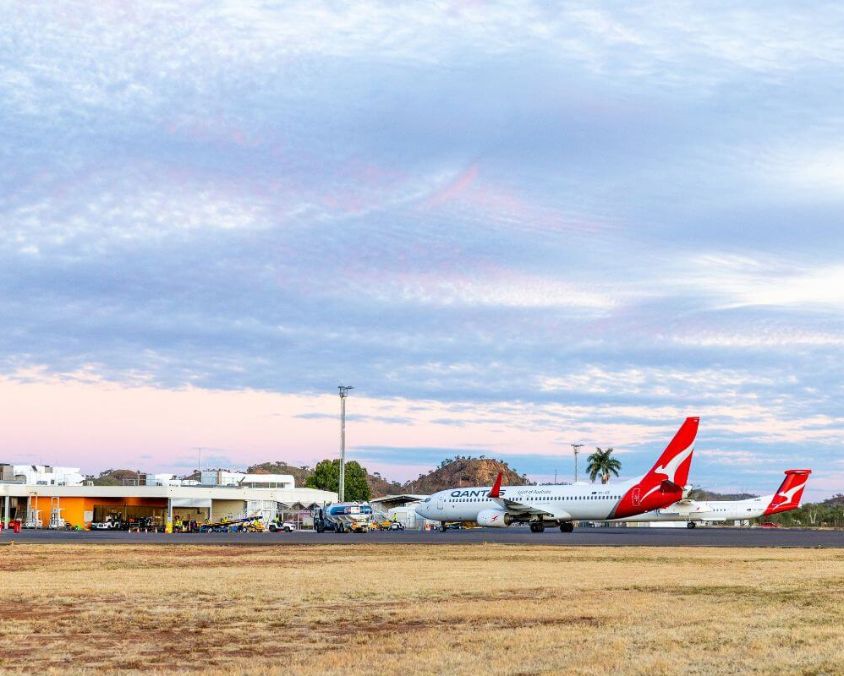 Two Qantas aircraft on the tarmac at Mount Isa Airport with terminal building and ground service vehicles in the background under a partly cloudy sky.