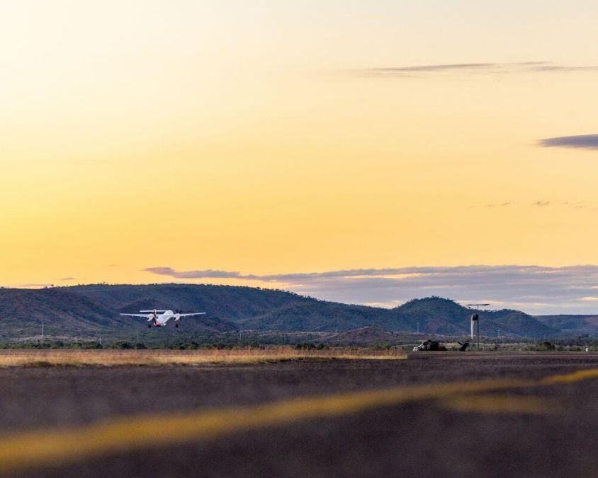 Small aircraft taking off or landing at Mount Isa Airport during sunrise or sunset, with hills and soft orange sky in the background.