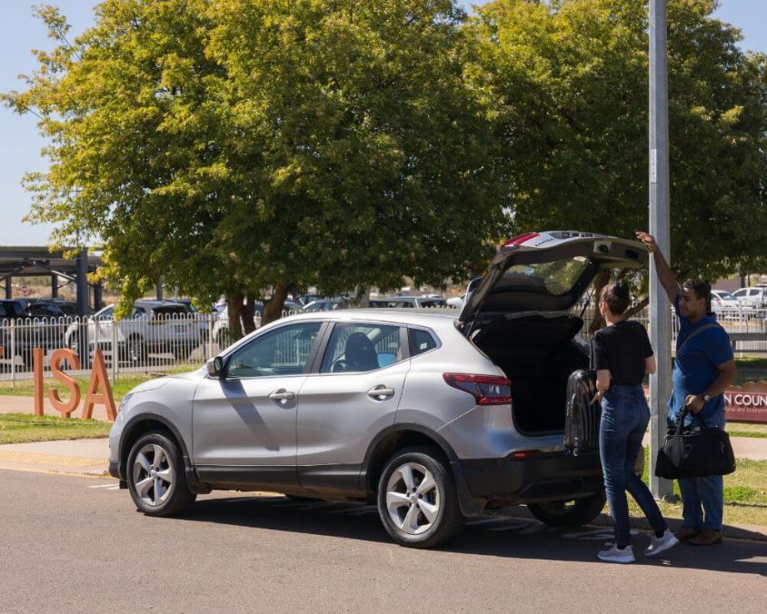Two people loading luggage into the boot of a silver SUV near car rental signage outside Mount Isa Airport.