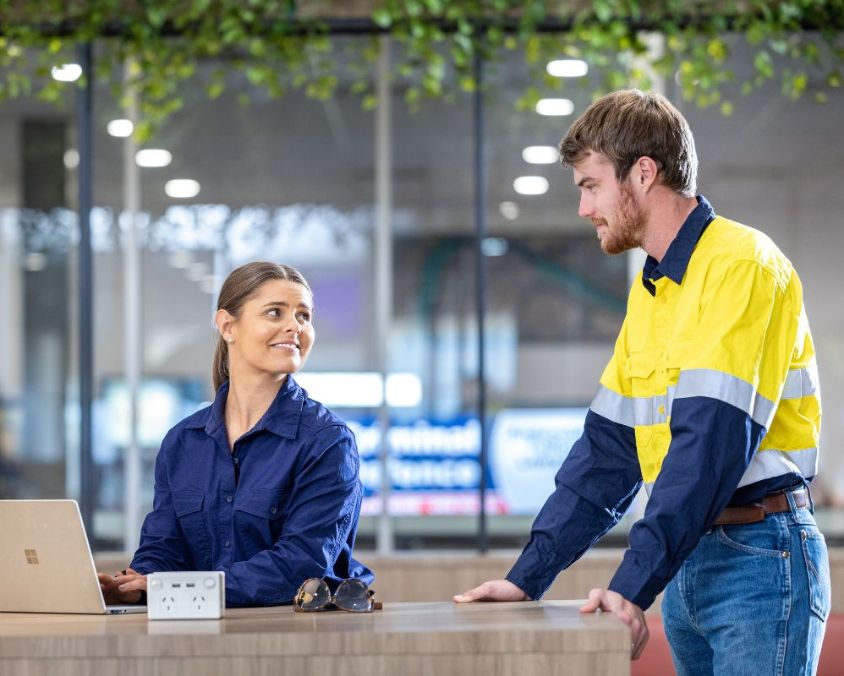 Two individuals interacting at a desk in the terminal of Mount Isa Airport, with a laptop and greenery visible in a modern office-style environment.