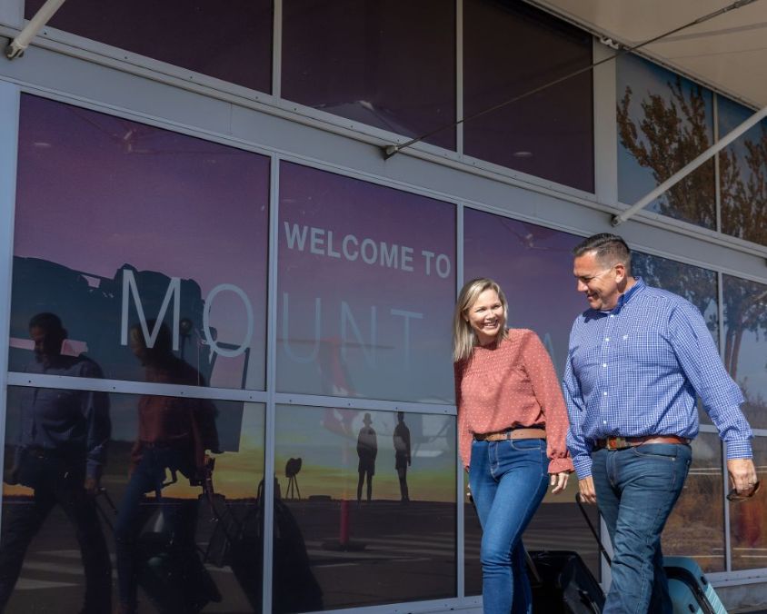 Two people walking outside the Mount Isa Airport terminal, with a large 'Welcome to Mount Isa' sign visible on the glass façade.