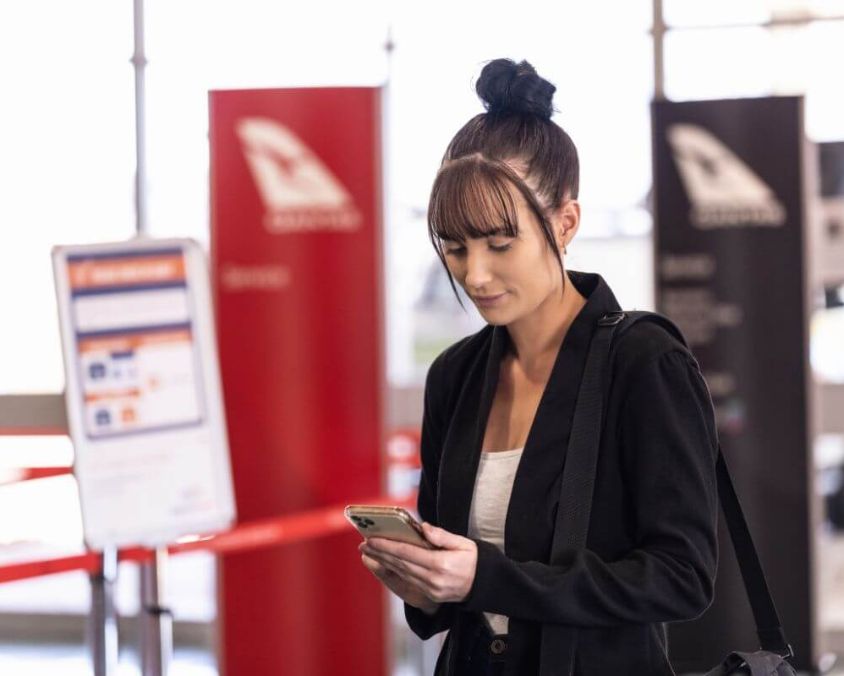 Person standing inside Mount Isa Airport terminal near Qantas signage, checking a smartphone with travel information displays in the background.